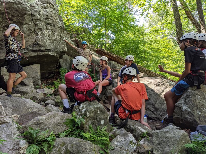 A group of young people wearing helmets are gathered on a rocky, wooded hillside. Some are sitting on the rocks, while others are standing. They appear to be preparing for or taking a break from a climbing or outdoor adventure activity. The scene is set in a natural environment with trees and large rock formations.